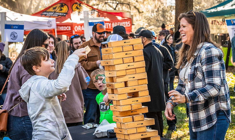 Boy and woman enjoying a Jenga game at an outdoor event, surrounded by people. Sunny setting with colorful tents in the background. Orlando Science Center's Science On Tap Beer Festival