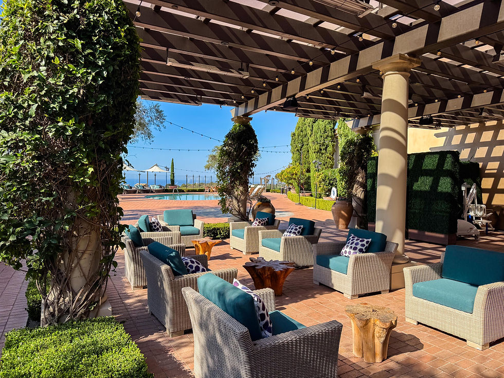wooden trellis above 9 wicker chairs with blue accent pillows and lots of landscaping with a pool and sun umbrellas in the back