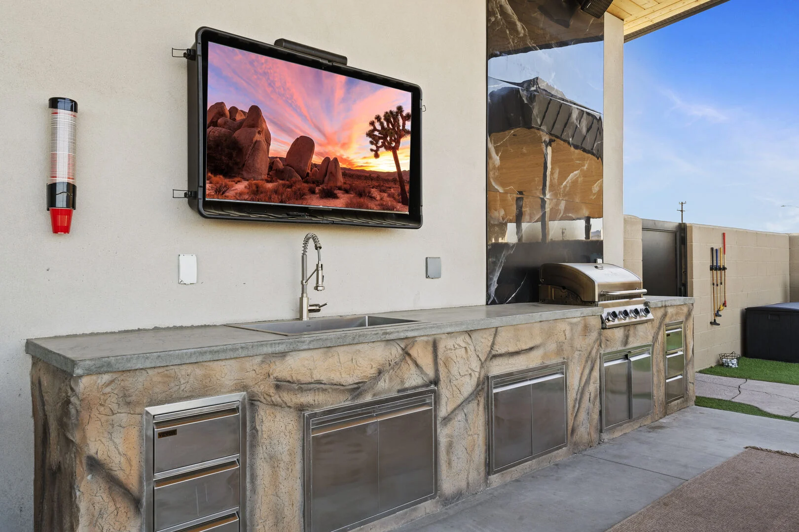 outdoor kitchen with stainless steel appliances and an outdoor tv on the wall for hosting and entertaining 