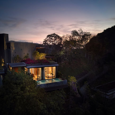 shadowed landscape illuminated by a pool and floor to ceiling glass windows looking in to a pool side villa with the twilight sky above