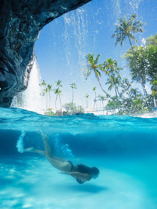 over under water photo with a woman swimming underwater below cave with a waterfall and palm trees