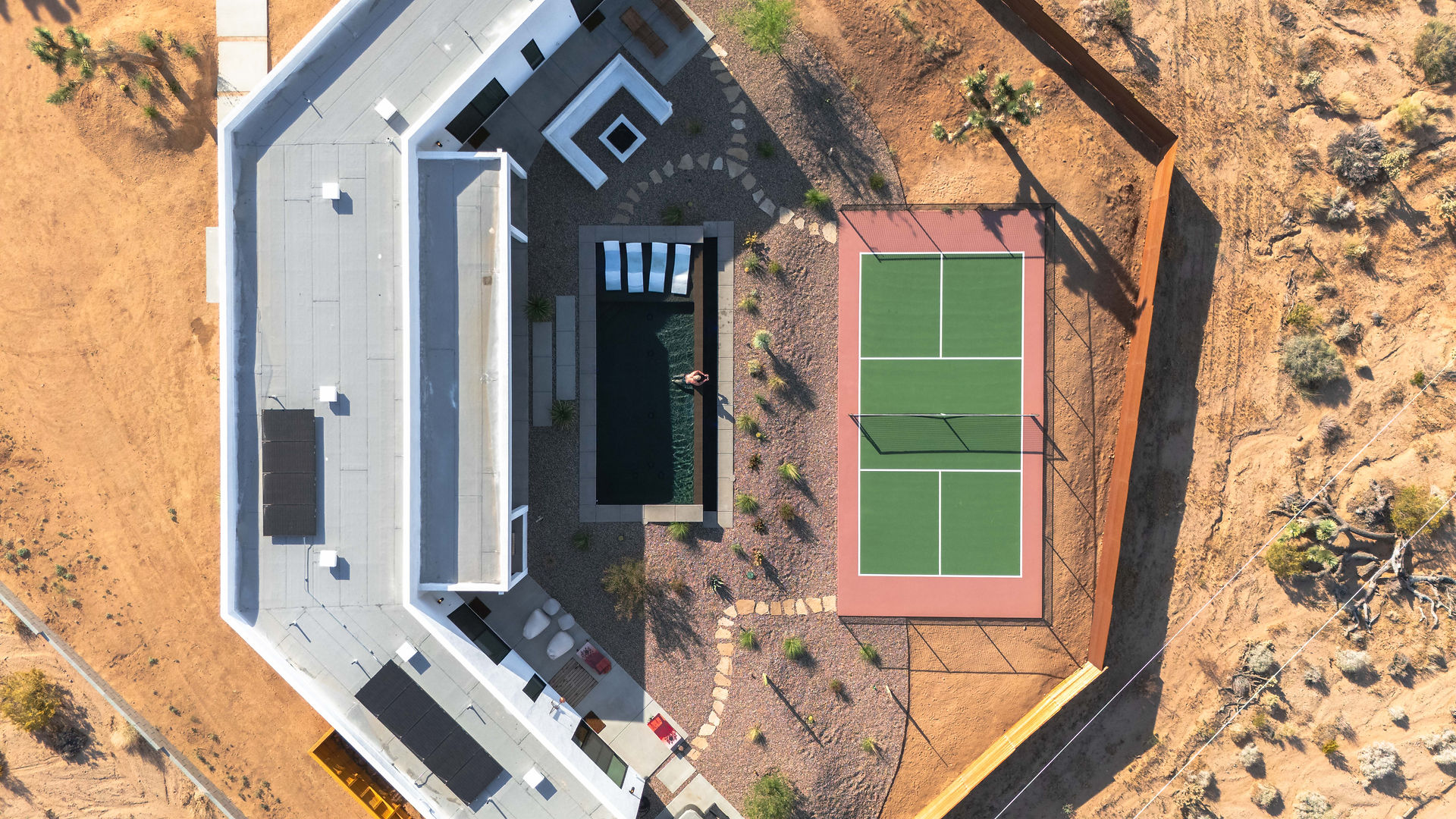 aerial view above a crescent shaped home featuring a pool and pickle ball court in the desert 