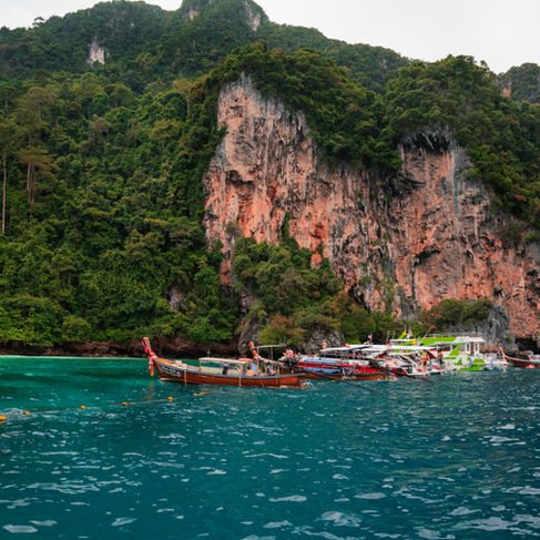 multiple Thai boats docked in the blue ocean waters with steep orange cliffs and hillside jungles in the background