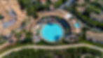 Aerial view looking down at the massive circular pool surrounded by lounge chairs and palm trees and a walking path around a resort with adjacent buildings and cars nestled in-between well manicured landscaped areas 