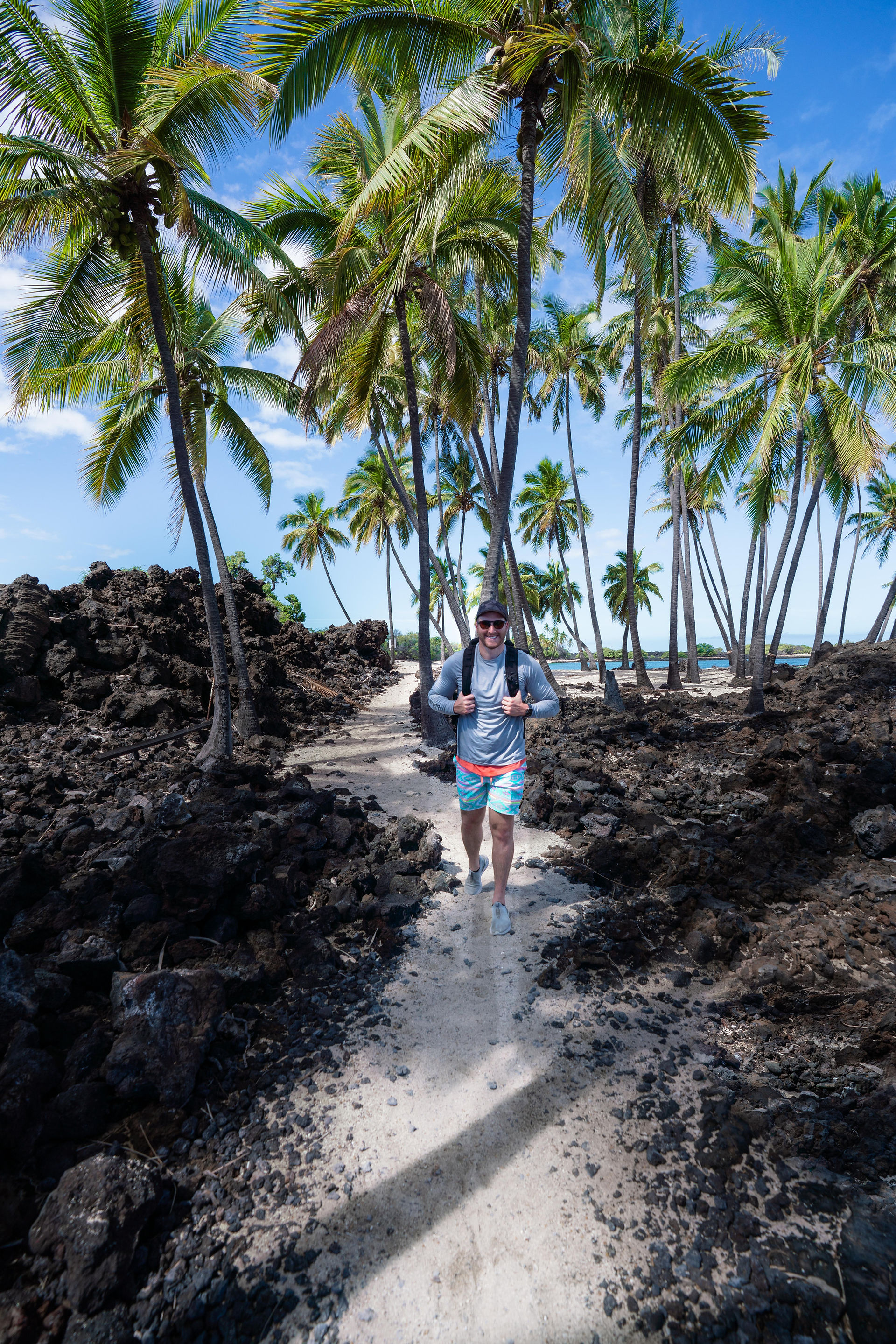 man hiking along a white sand pathway between black lava rocks with a cluster of palm trees standing tall behind 