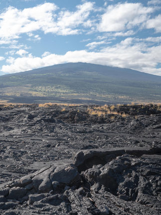 serpentine lava flow formations of black rock from the high mountain volcano in the distance under blue sky and clouds
