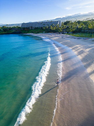 waves crashing on to the white sand beach with a man standing in the water with palm trees and a hotel tower in the distance