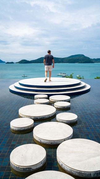 man in shorts and t shirt standing in the center of a large white deck with pavers on top of water leading towards the ocean and islands in the distance