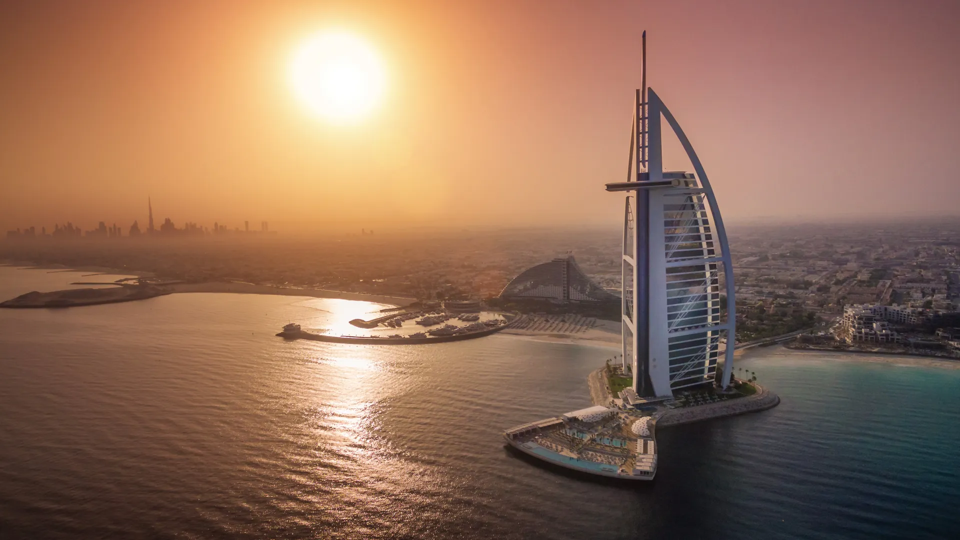 The Burj Arab structure in front of the Dubai city skyline underneath a sun setting 