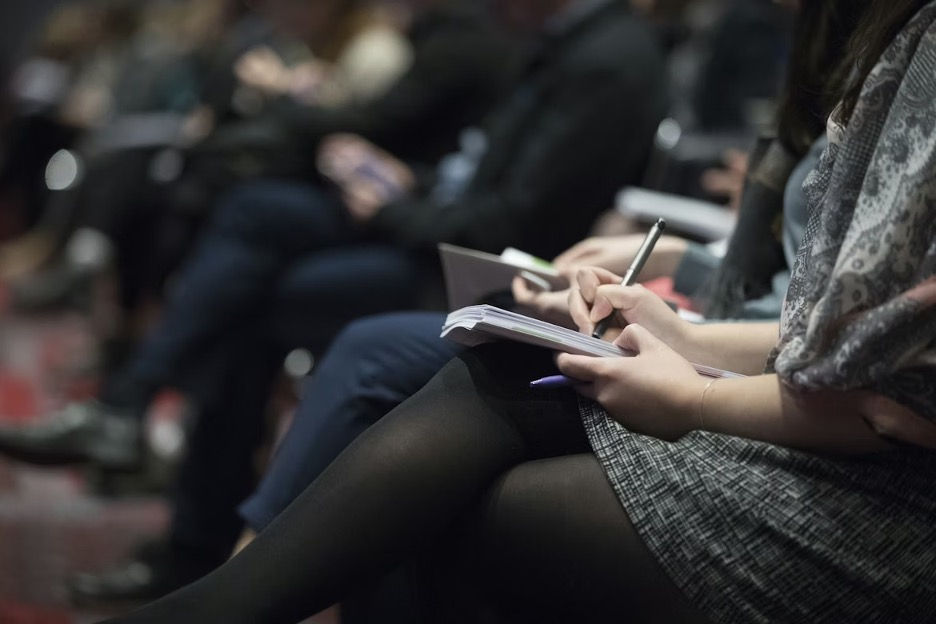 Audience members in a dim lecture hall take notes in notebooks during a presentation.