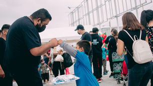 Man fist-bumps a boy in a blue shirt in a crowded outdoor area with metal railings.