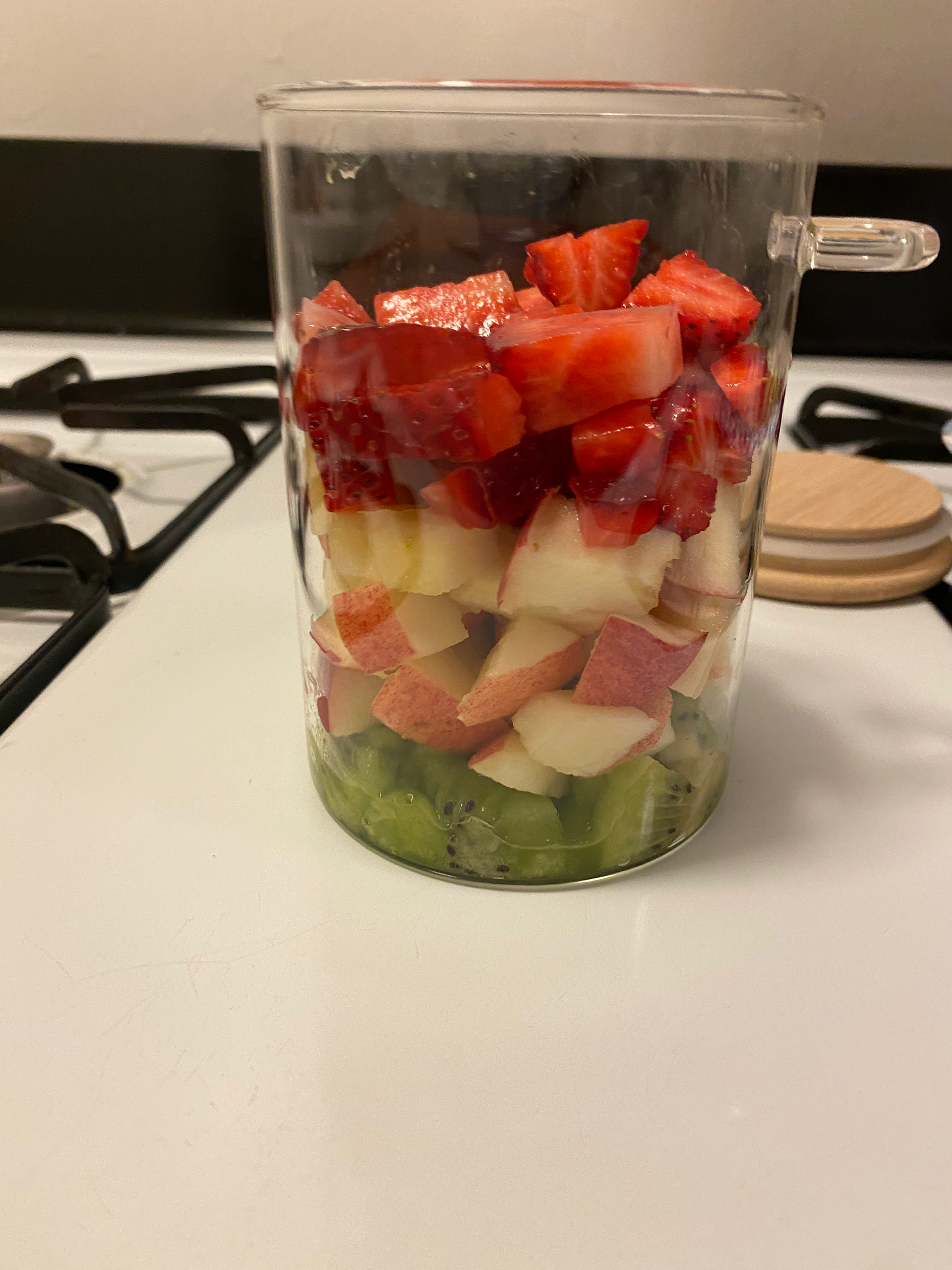Layered fruit salad in a glass cup with strawberries, apples, and kiwi. The cup is on a stove-top, creating a fresh, colorful scene.