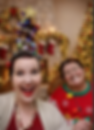 Two people smiling in a festive setting. One wears a tall, decorated Christmas tree hairstyle. Colorful lights and stockings in the background.