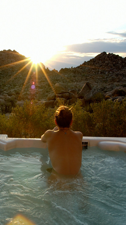 A person relaxes in a outdoor hot tub, looking out at a stunning desert sunset behind rocky mountains.