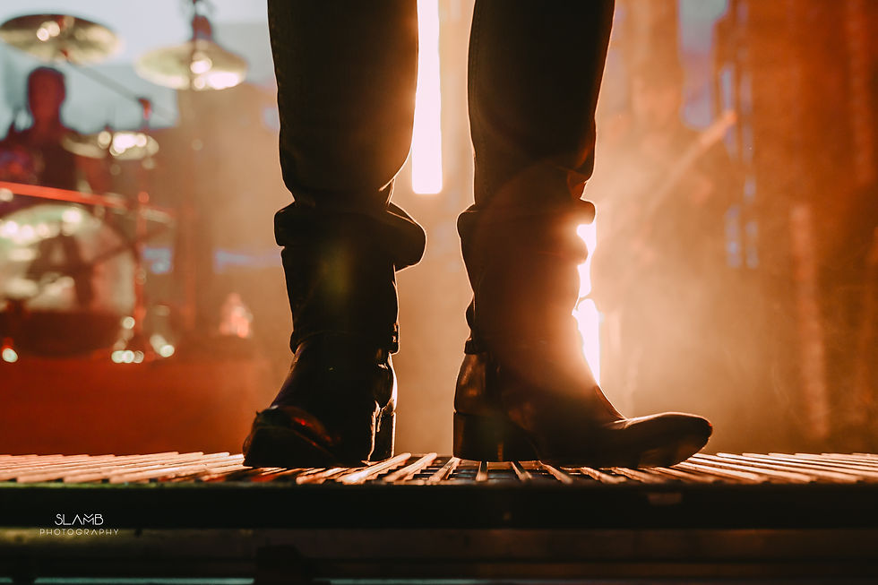 Two feet, wearing black boots, standing on a metal platform. There is an orange light beaming from behind them.