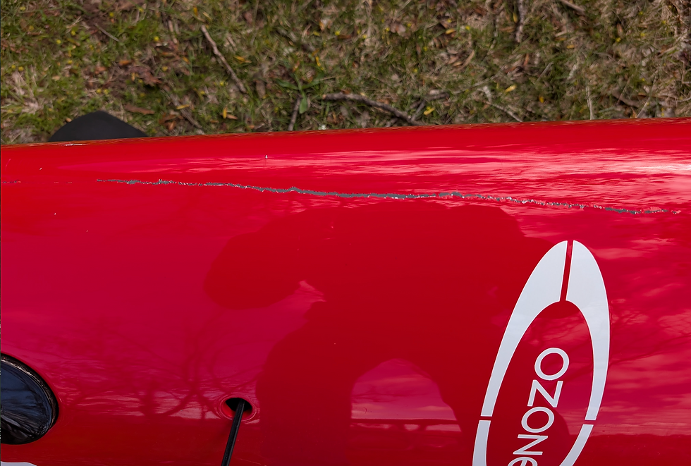 Red kayak with a scratch on the surface, "OZONE" text visible. Background shows grass and twigs. Shadow of a person reflected.