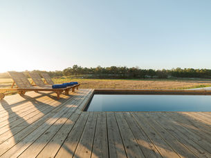 Wooden deck with three loungers and blue towels beside a reflective pool. Open field and trees in the background under a clear sky.