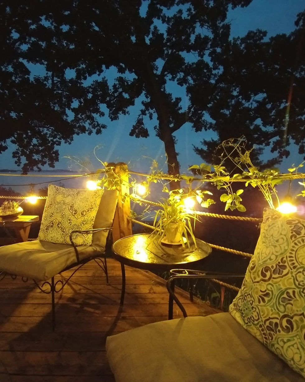 Cozy patio setting at dusk with string lights, two cushioned chairs, patterned pillows, a small table with a plant, and silhouetted trees.