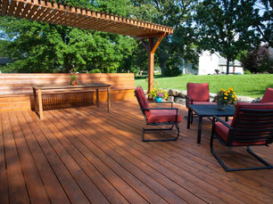 Wooden deck with a pergola, featuring a dining table, red-cushioned chairs, and colorful flowers. Green lawn and trees in the background.