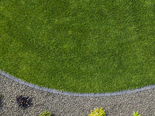 Circular green lawn bordered by bricks, adjacent to a gravel area with small plants. Vibrant greens and grays create a serene garden scene.
