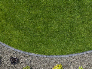 Curved green lawn bordered by dark stones, meets a gravel area with small, colorful plants. A serene, tidy garden scene.