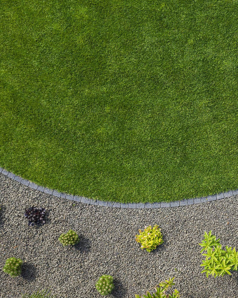 Curved green lawn bordered by dark stones, meets a gravel area with small, colorful plants. A serene, tidy garden scene.