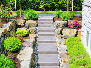 Stone steps lead through a lush garden with vibrant flowers and shrubs, framed by stone walls and trees, creating a serene outdoor scene.
