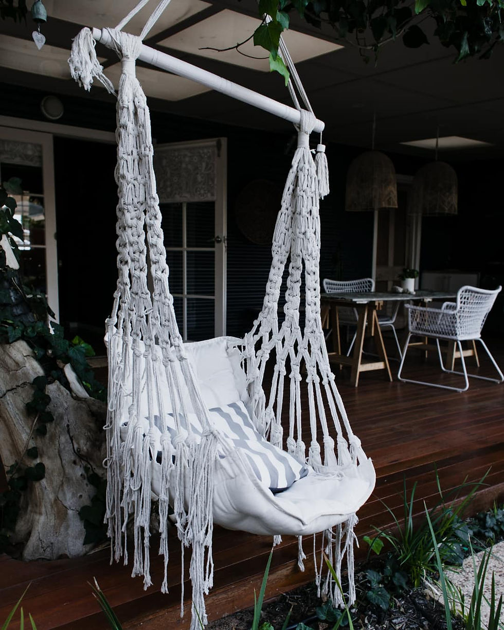 White macrame hammock chair with fringe hangs on a wooden porch. A table with chairs is in the background, creating a relaxed outdoor vibe.