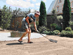 Man in a hat rakes soil in a garden, surrounded by tall evergreens and a brick house. Bright, sunny day with blue sky and clouds.