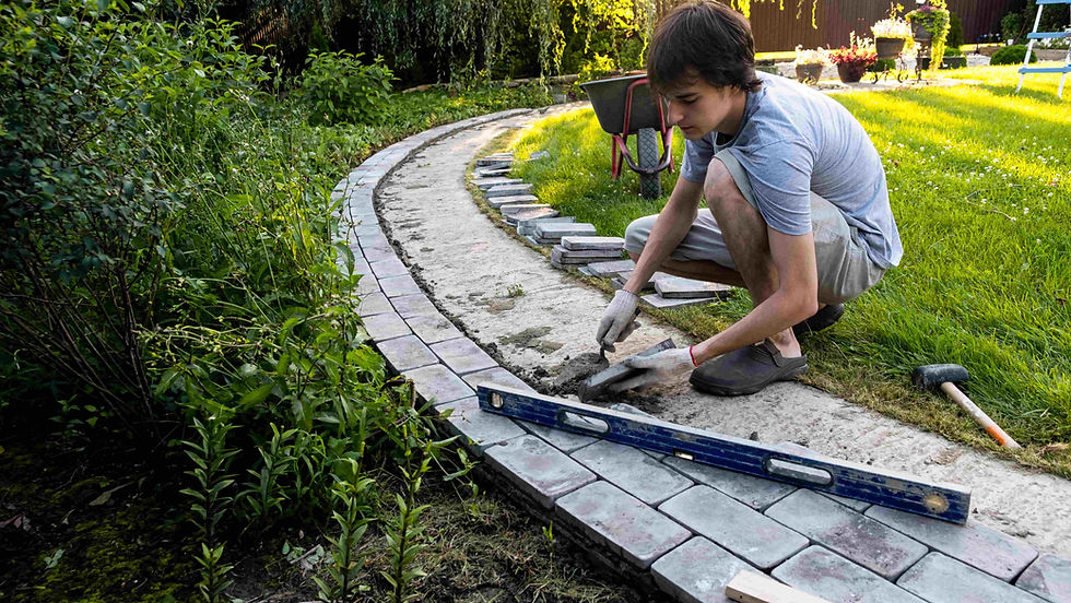 A person laying bricks on a curved garden path surrounded by greenery. Nearby are tools, a wheelbarrow, and sunlight filtering through trees.