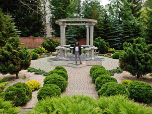 A person stands smiling in a lush garden with a circular stone pergola. Green shrubs and brick pathways surround them.