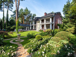Elegant brick mansion with white trim nestled in lush garden, tall trees surrounding, clear blue sky. Pathway leads to a birdbath. Serene mood.