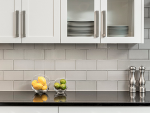 Modern kitchen with white cabinets, glass doors showing plates, black countertop with lemons, limes in bowls, silver shakers, gray tile backsplash.