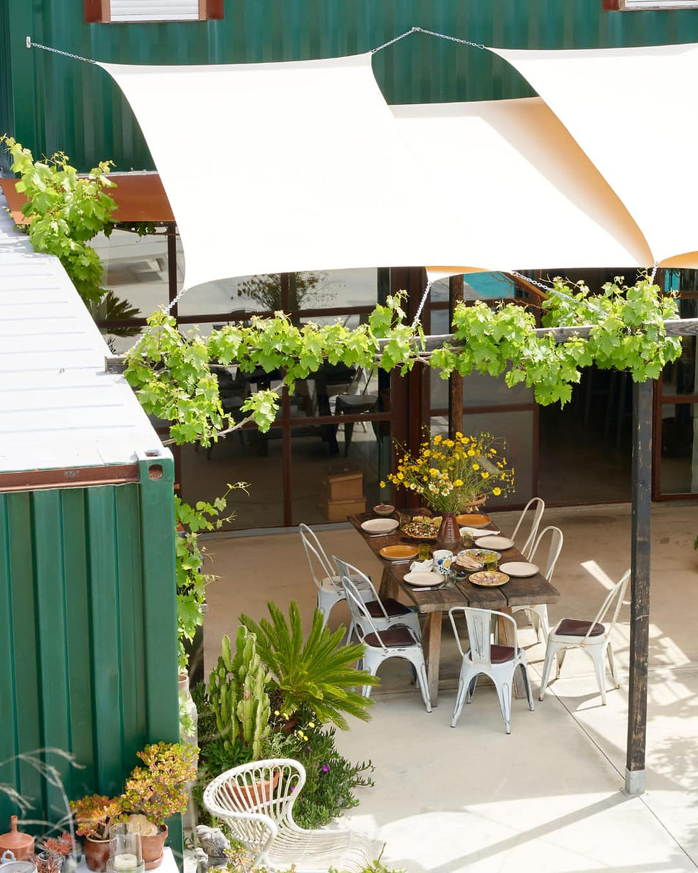 Wooden table with plates of food under a white canopy. Surrounded by chairs, plants, and green panels, creating a relaxed outdoor vibe.