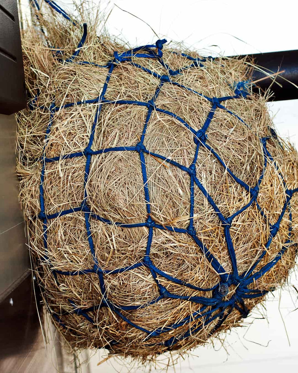 Hay bale enclosed in a blue net hangs on a metal bar. The background is white, creating a clean and organized appearance.