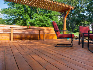 Wooden deck with red-cushioned chairs, a table, and a pergola. Green trees in the background, under a clear blue sky. Relaxing mood.