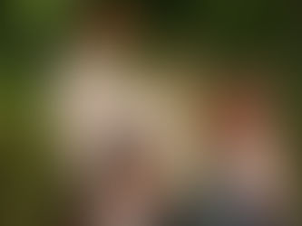 Family of five stands in tall grass, looking thoughtful. Adult in red cap, woman hugging child. Trees in background, natural setting.