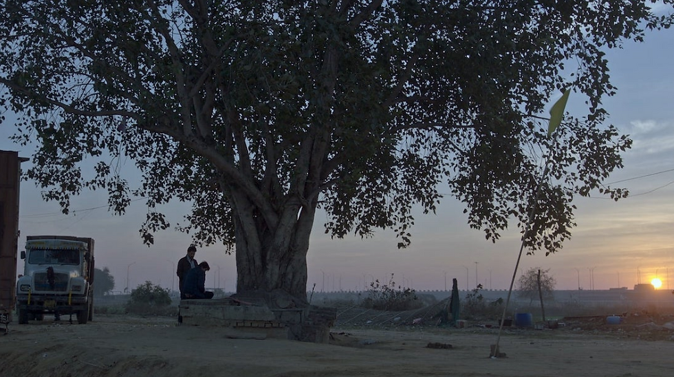 Two people sit under a large tree at sunset near a parked truck, with a rural landscape in the background creating a serene mood.