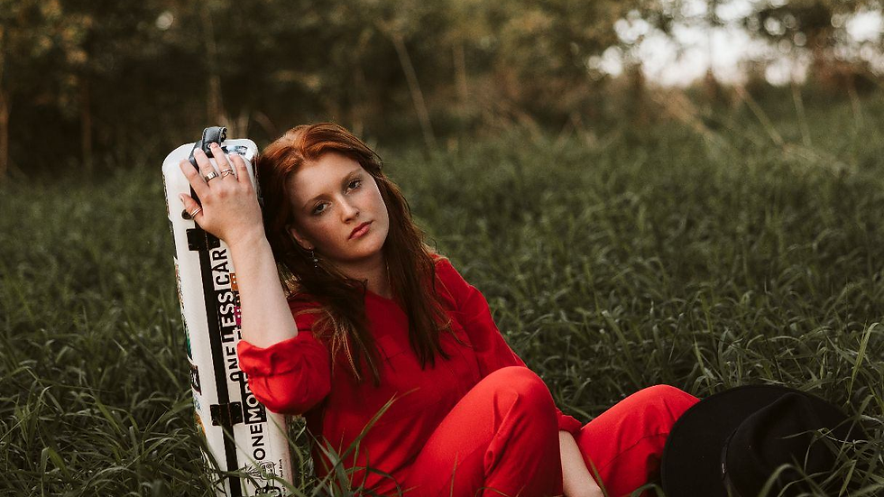 Woman in red sits in grass, holding a skateboard with "One More, One Less Car." She looks thoughtful, surrounded by trees.
