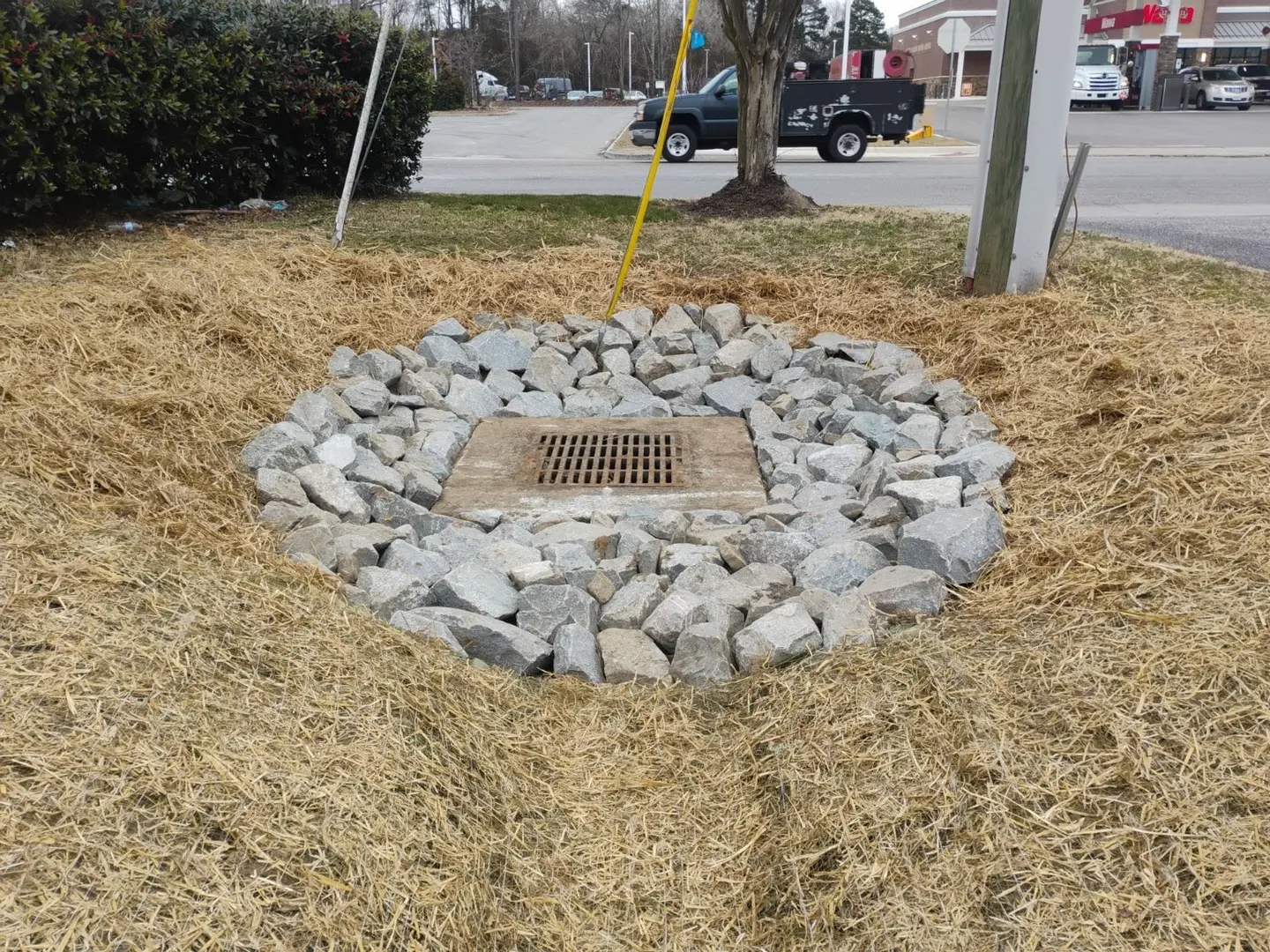 A drainage inlet surrounded by riprap and straw-covered ground.