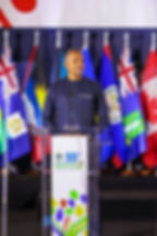 Tall dark-skinned man standing at a podium with many Caribbean flags in the background.
