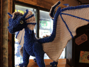 A blue and grey crochet dragon with bright blue-green eyes.