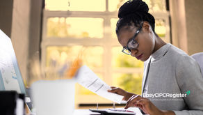 A person in a suit working at a desk, reviewing documents and using a calculator, with a computer screen and a window in the background.