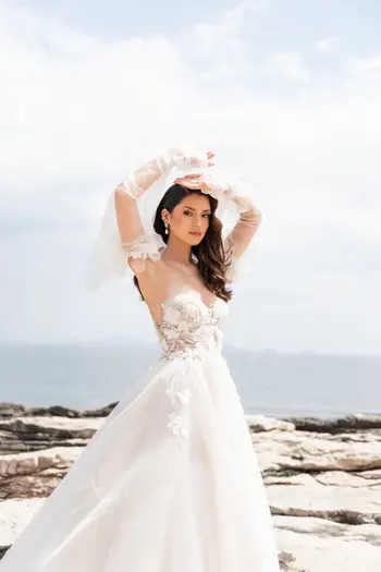 Hermosa novia con vestido de novia blanco, guantes, brazos en alto, costa rocosa y mar.