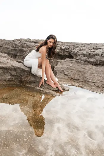 Mujer con un vestido blanco sentada cerca de un charco en unas rocas.