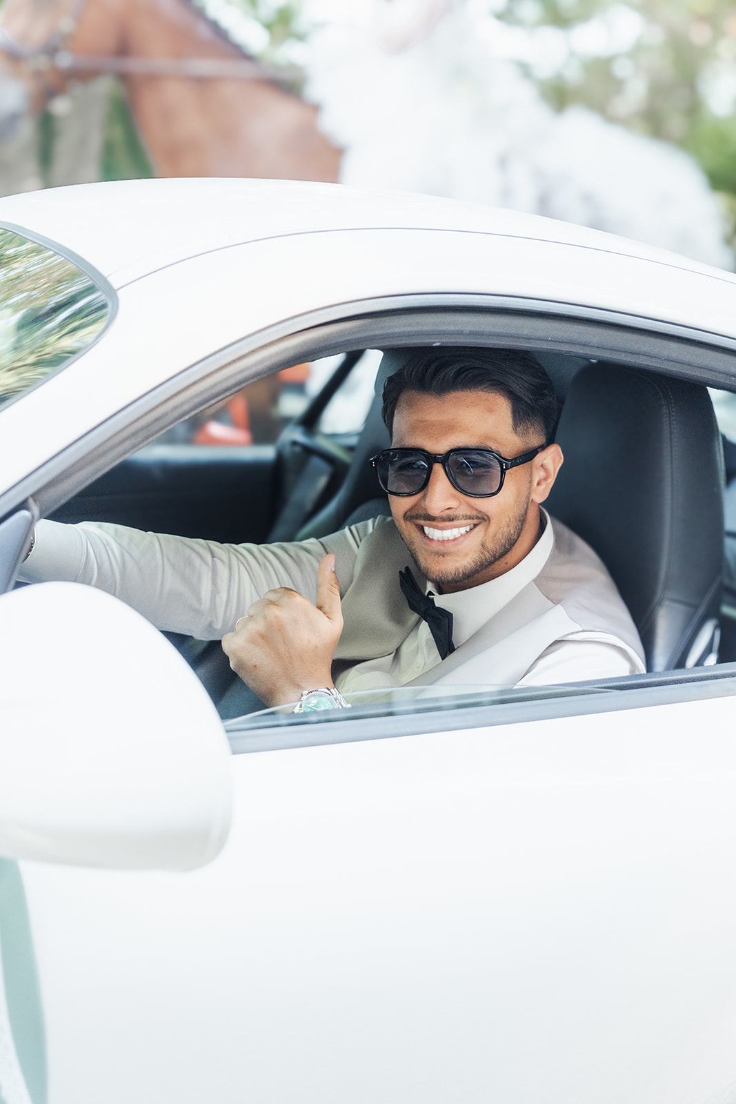 Smiling man in sunglasses in a white car, giving a thumbs-up gesture.