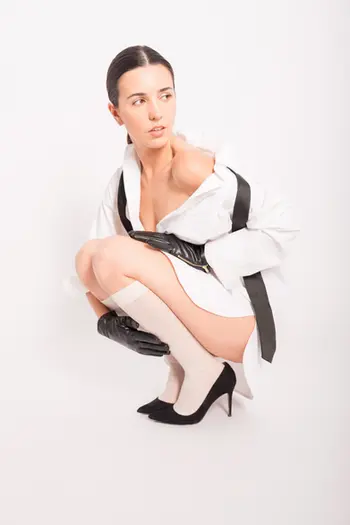 Una donna accovacciata, con indosso una camicia bianca, posa con sicurezza su uno sfondo bianco, guardando verso sinistra.