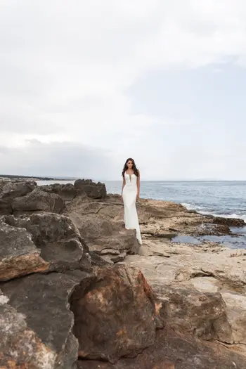 Mujer con vestido blanco posando sobre rocas frente al océano, al aire libre.