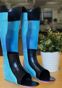 Two bright blue AFOs fitted with black TPU inserts from the front beside a plant.