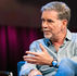 Reed Hastings speaks during a TED on-stage interview, seated and gesturing while wearing a headset mic.